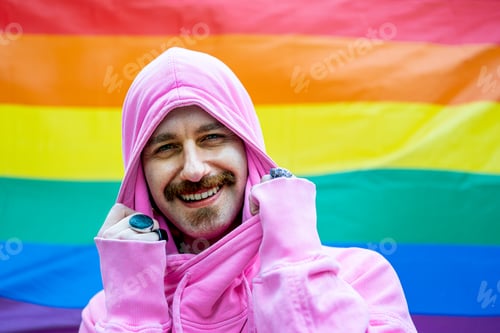 Preview: happy gay man smiling and looking at the camera, rainbow flag on background