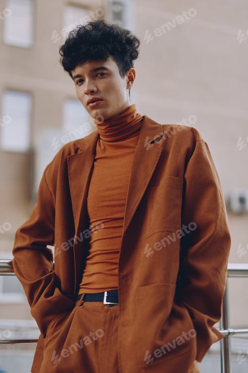 Preview: A stylish young man with curly hair in a brown suit and turtleneck, standing casually with a modern