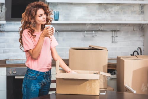 Preview: attractive woman holding cup of tea and unpacking cardboard box at new home