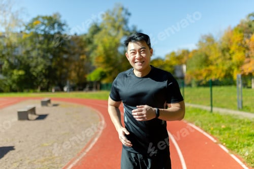 Preview: Portrait of a young Asian sportsman runner in black sportswear running in the stadium and smiling