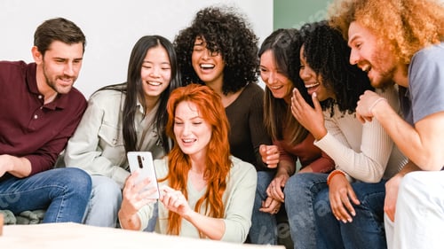 Preview: Happy woman showing smartphone to cheerful diverse friends in living room at home