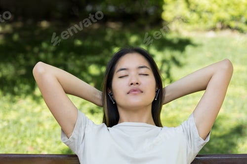 Preview: Young Asian woman on bench outdoors, listening to music with eyes closed, enjoying nature