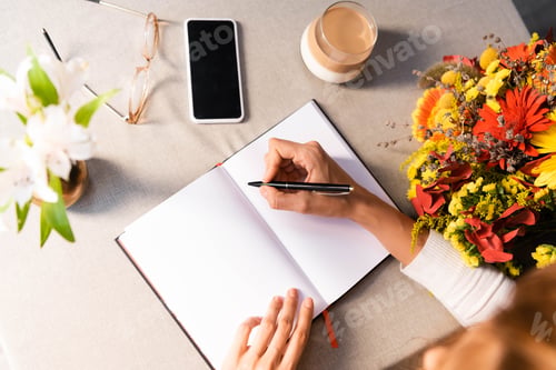 Preview: cropped view of woman writing in notepad in cafe with coffee, smartphone and flowers