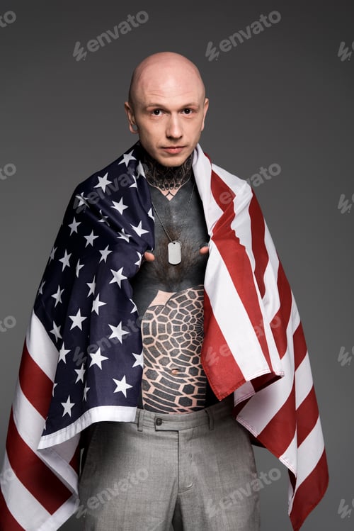 Preview: bald shirtless tattooed man standing with us flag and looking at camera isolated on grey
