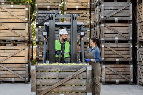 Preview: Happy young bearded guy driver in helmet in forklift truck with box full green apples