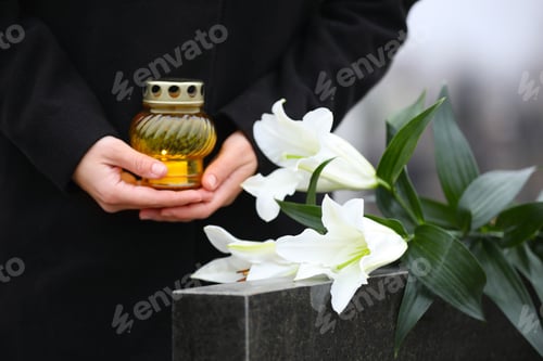 Preview: Woman with candle and white lilies near black granite tombstone outdoors, closeup. Funeral ceremony