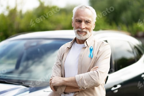 Preview: Senior man wearing blue ribbon standing with arms crossed in front of his car