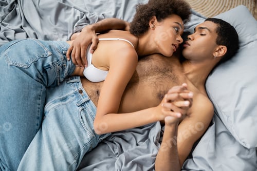 Preview: passionate african american couple in jeans holding hands and looking at each other on bed