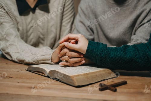Preview: Group of Christians sit together and pray around a wooden table with blurred open Bible pages