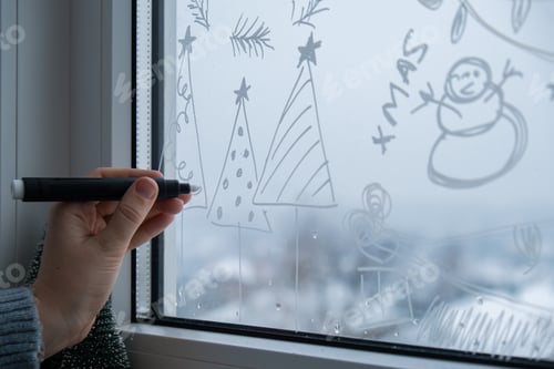 Preview: Creative enthusiastic unrecognizable woman in sweater paint window with white marker. Christmas tree
