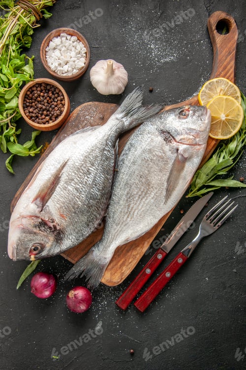 Preview: top view raw sea fish on chopping board fork and knife onions sea salt and black pepper in small