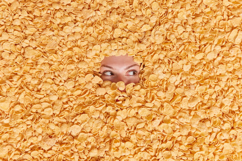 Preview: Top view of young woman fully covered in cereals focused away buried in cornflakes makes creative sh