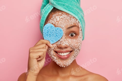 Preview: Headshot of cheerful young woman covers one eye with heart shaped cosmetic sponge, removes makeup, h
