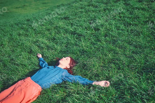 Preview: Teenage girl with red hair lying on green grass with arms spread.