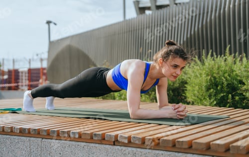 Preview: woman in a blue sports top performs a plank exercise on outdoor mat, surrounded by urban greenery