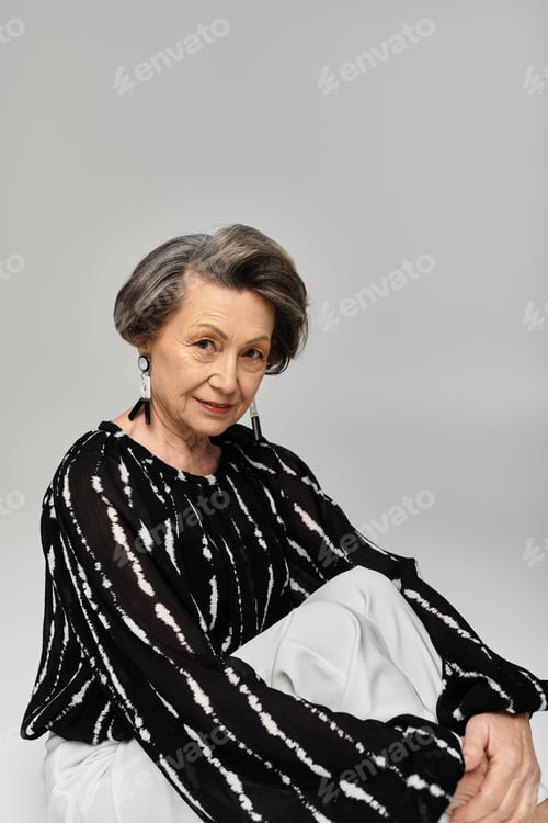Preview: A beautiful woman in her 60s poses gracefully on a grey backdrop, displaying her elegant attire.