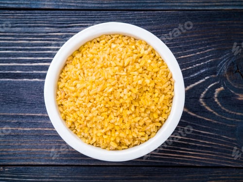 Preview: White bowl with raw bulgur on dark brown wooden background. Top view.