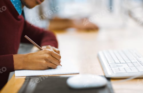 Preview: Shot of a unrecognizable student writing down notes while working on a computer in a library