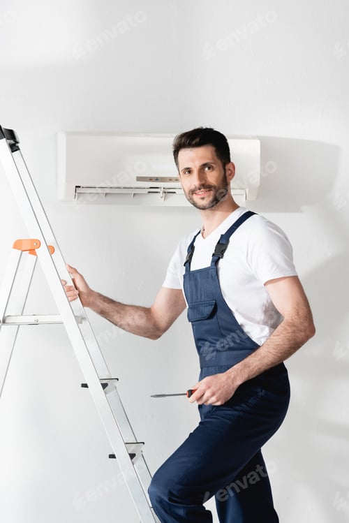 Preview: smiling repairman holding screwdriver while standing on stepladder near air conditioner