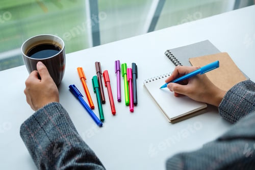 Preview: Closeup image of a woman writing on a blank notebook with colored pens while drinking coffee