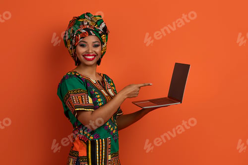 Preview: Happy black woman in african costume pointing at laptop screen