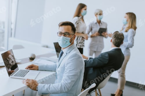 Preview: Young business man with medical protective mask works on a laptop