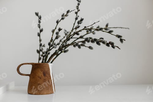 Preview: Willow branches in stylish rustic vase on wooden table. Simple Easter rural minimal aesthetics