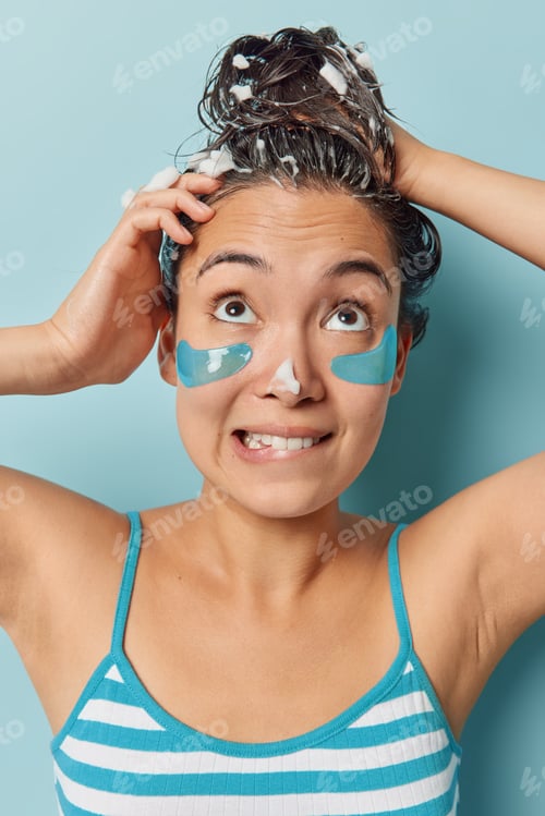Preview: Vertical shot of brunette Asian woman washes head with shampoo applies beauty patches under eyes to
