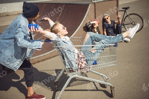Preview: Boy carrying happy girl in shopping cart while friends sitting near ramp at skateboard park