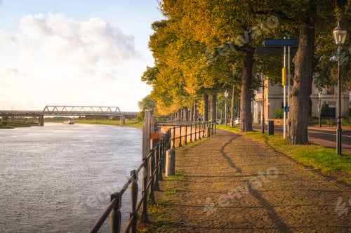 Preview: Scenic view of a walkway along a river lined with green trees at golden hour
