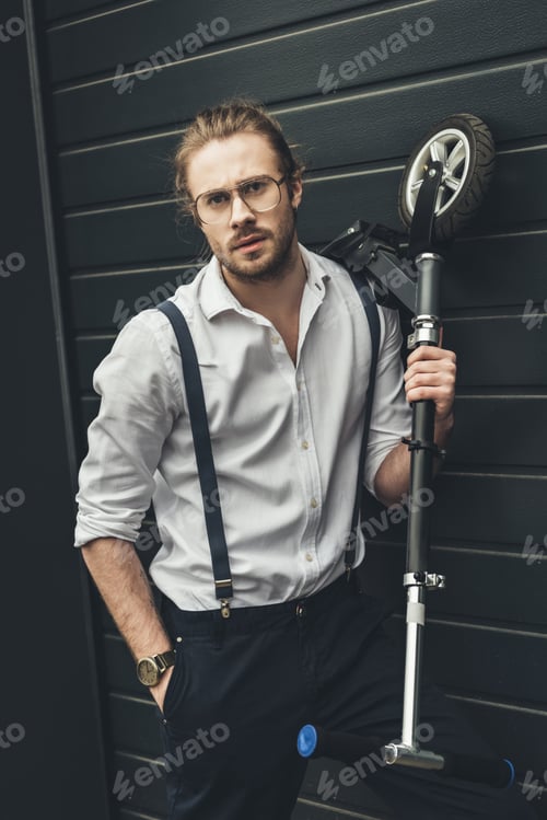 Preview: Handsome stylish young man in spectacles posing with scooter and looking at camera