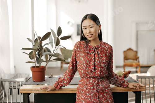 Vista previa: Happy Asian brunette woman in red stylish floral dress smiles, looks into camera and leans on woode