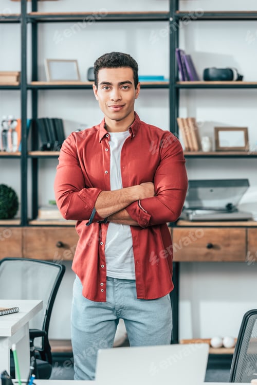Preview: Joyful young man in red shirt standing with crossed arms at workplace