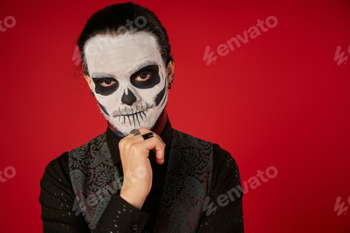 Preview: man in spooky skeleton makeup holding hand near chin and looking at camera on red, Day of Dead