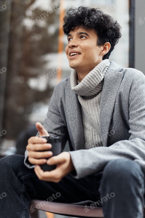 Preview: Young man sitting on bench with hands in pockets, coffee cup in front of face