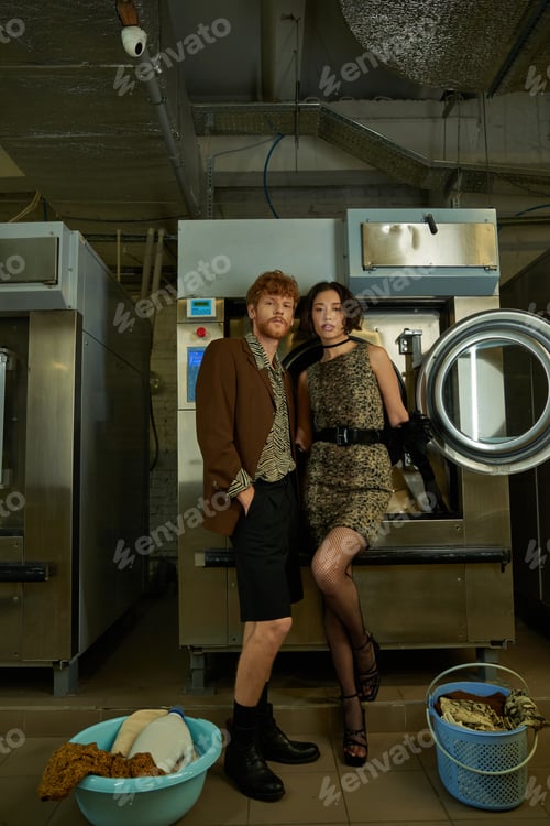 Preview: fashionable young interracial couple looking at camera near clothes in coin laundry