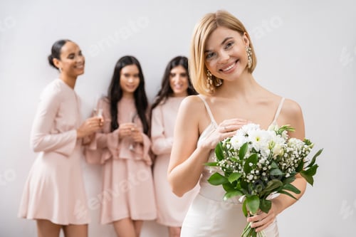 Preview: attractive bride in wedding dress holding bouquet, standing near blurred interracial bridesmaids