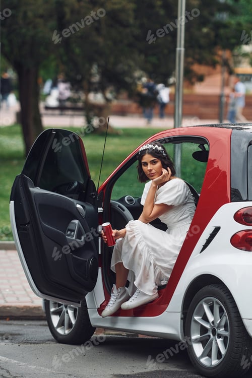 Preview: Sitting in white and red colored car. Bride in dress is outdoors before her wedding begins