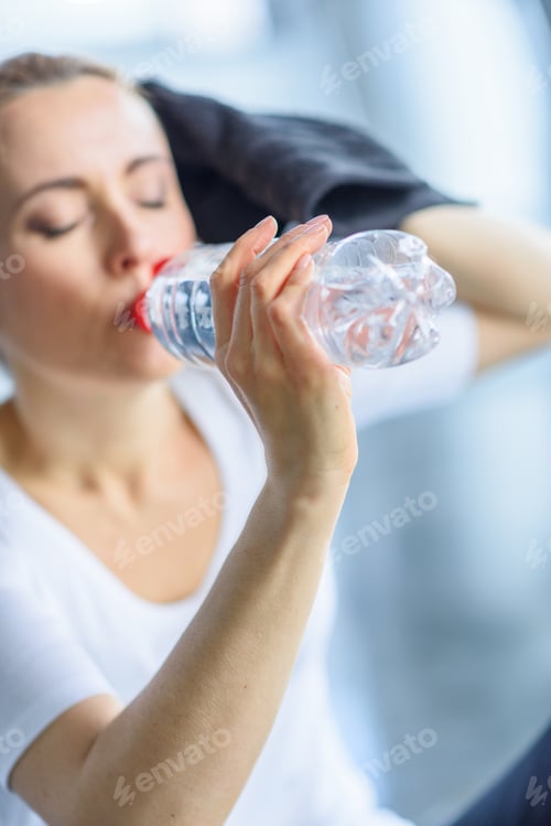 Preview: tired sportswoman drinking water from sport bottle in sports center