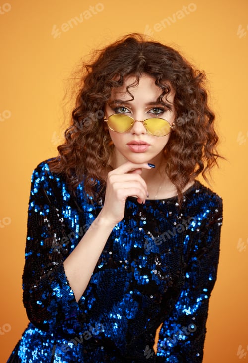 Preview: Vertical portrait of adorable curly-haired girl put her finger to her chin on orange background