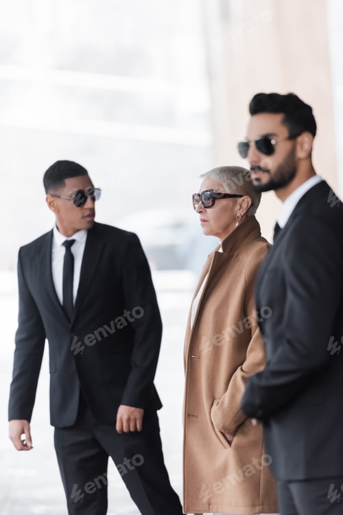Preview: selective focus of wealthy businesswoman with hand in pocket near private interracial bodyguards