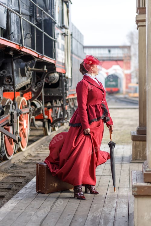 Preview: A beautiful girl in a burgundy suit and a hat stands with an umbrella near an old steam locomotive.
