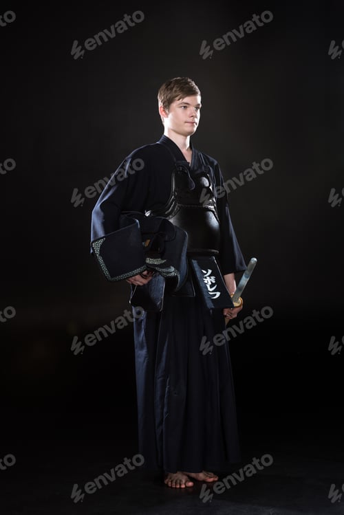 Preview: Full length view of young man holding kendo helmet and bamboo sword on black