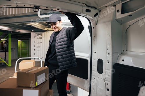 Preview: Confident courier leaning on van looking on cardboard boxes