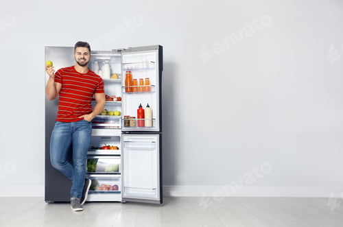 Preview: Happy young man with apple near open refrigerator indoors, space for text