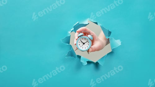 Preview: Hand holds alarm clock through a paper hole. Time management and deadline concept