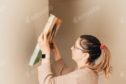 Preview: Young millennial mother at home hanging up picture of daughter against beige colored wall