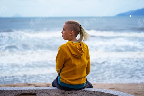 Preview: Lonely teenager looking to sea sitting on the waterfront. Rear view of teenager against seascape.