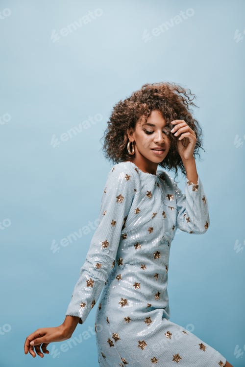 Preview: African girl with fluffy brunette hair in long sleeve blue dress and modern gold earrings posing on