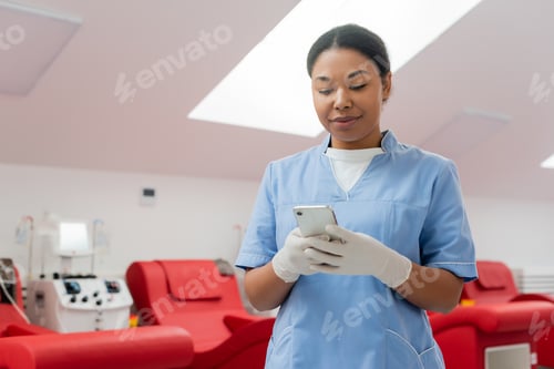 Preview: positive multiracial doctor in blue uniform and latex gloves messaging on smartphone near